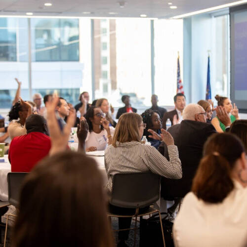 A group of people sit in a room. Some raise their hands as they face a white board. The US flag and GVSU stand beside